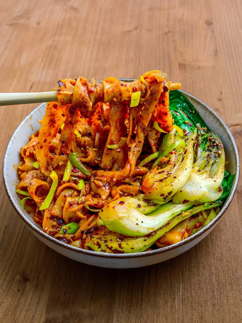 Nouilles biang biang sur fnd de bois dans un bol avec du pak choi soulevées par des baguettes