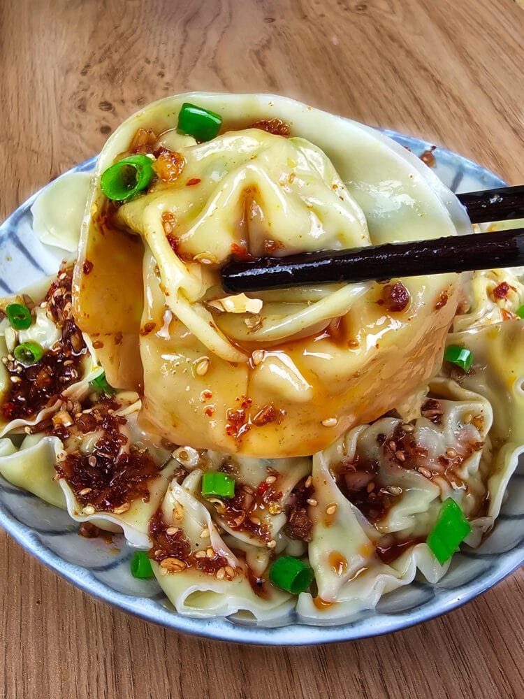 wonton enrobé de sauce sichuanaise pimentée tenu par des baguettes devant une assiette bleutée remplie de wonton sur fond de bois