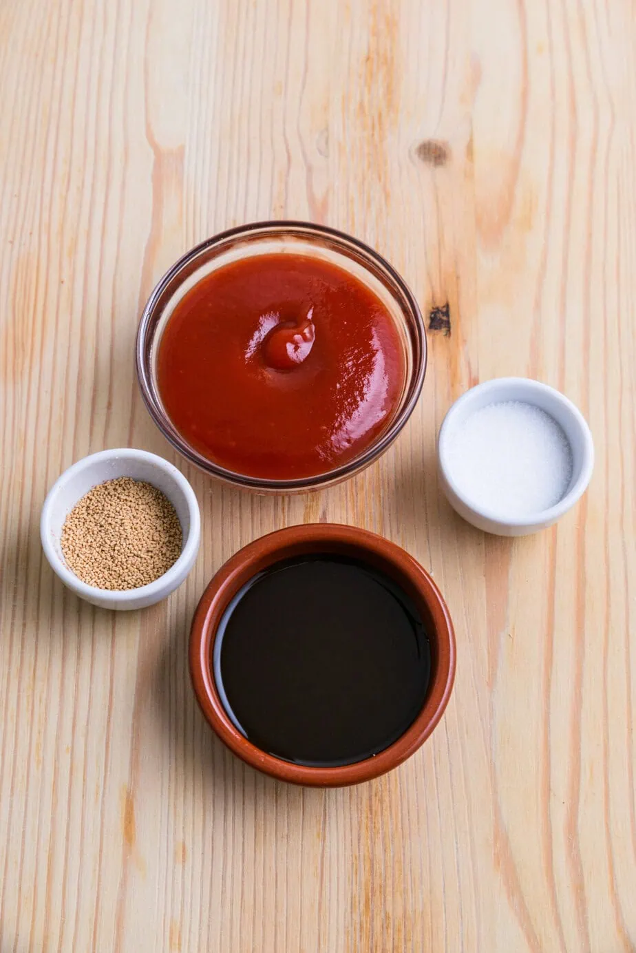 Ingrédients pour sauce bulldog maison dans 4 petits bols sur un fond en bois clair. Le ketchup rouge est au-dessus, la sauce huitre noire en bas, le dashi en granules à gauche et le sucre à droite