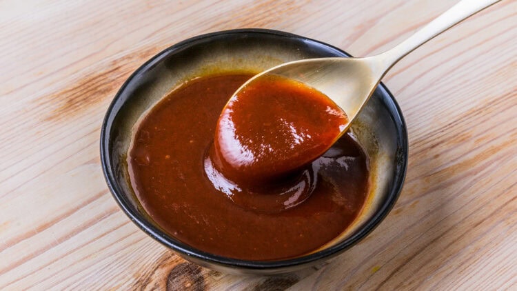 Sauce tonkatsu rougeâtre dans un bol noir et jaune sur fond de bois, avec une cuillère en bois clair soulevant un peu de sauce bulldog