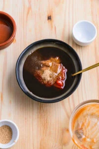 Ingrédients pour la sauce tonkatsu maison mélangés dans un bol sur un fond de bois clair, avec des bols à condiments vides de par et d'autre