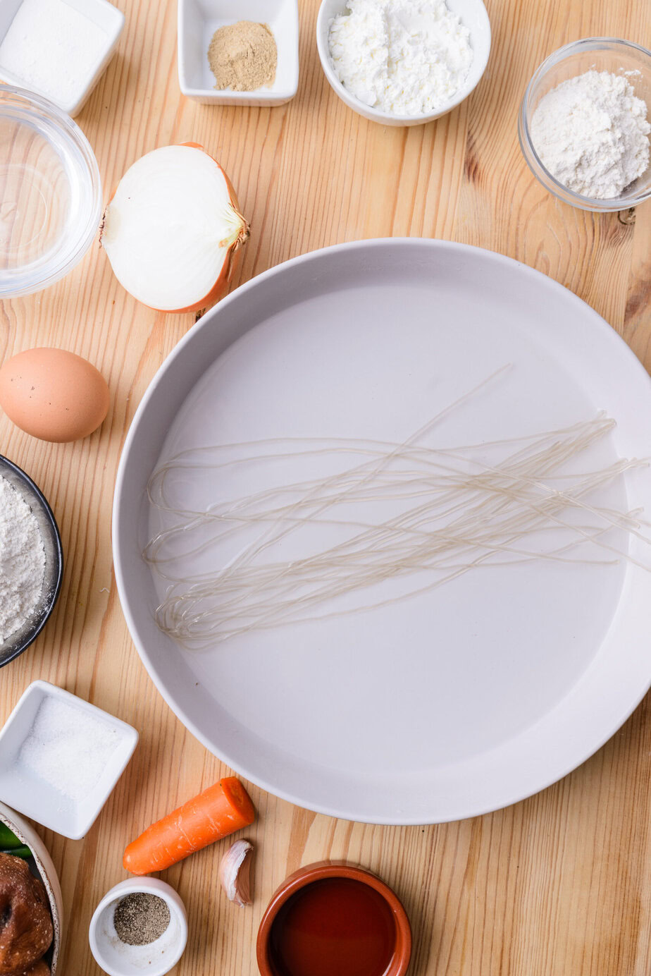 Ingrédients variés sur une table en bois, dont un œuf, une demi-oignon, des vermicelles et des épices.