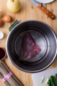 Un morceau de viande crue dans une casserole, entouré d’ingrédients sur une table en bois.