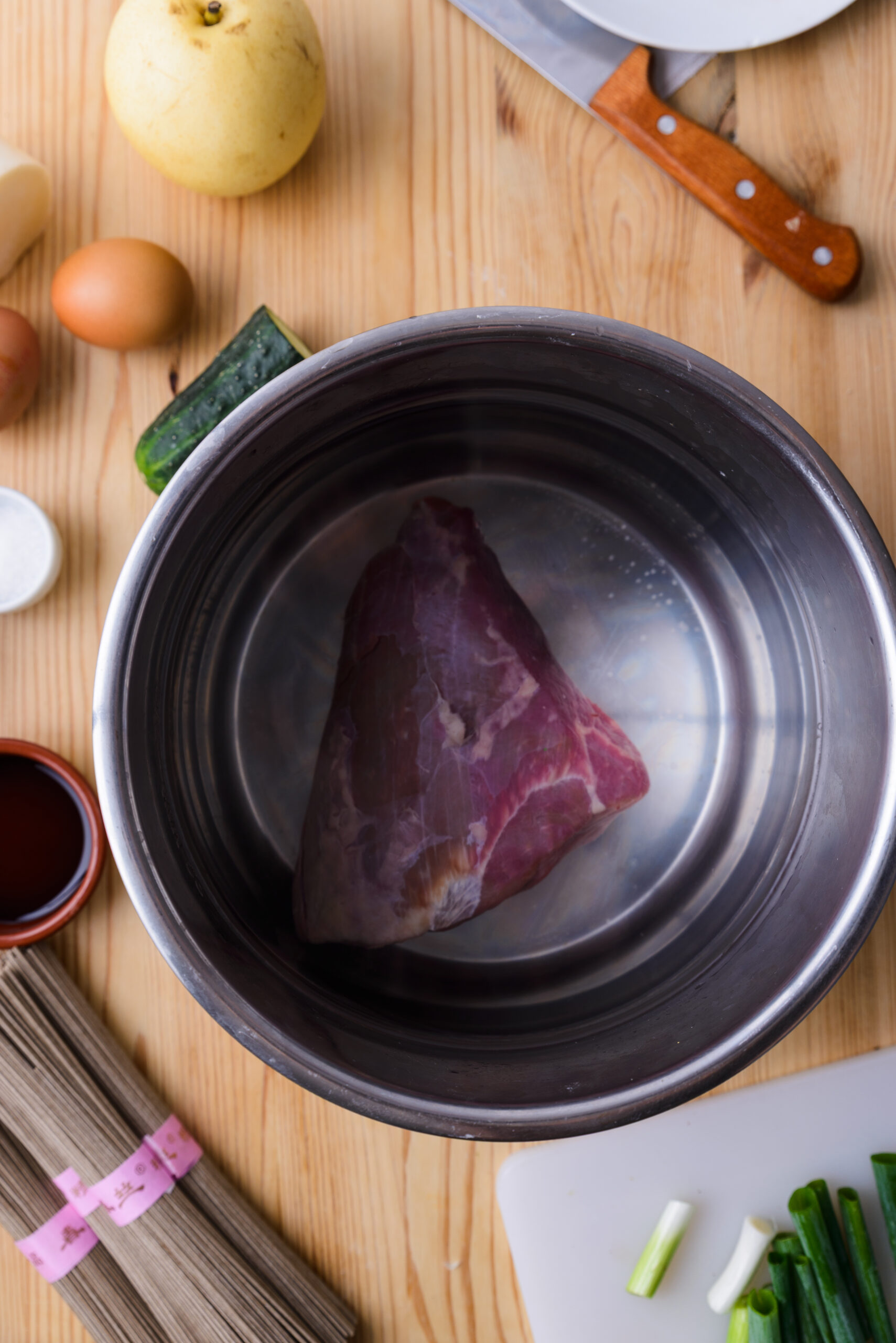 Un morceau de viande crue dans une casserole, entouré d’ingrédients sur une table en bois.