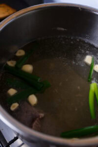 Bouillon en cuisson dans une casserole avec des morceaux d’ail et des oignons verts flottant à la surface.