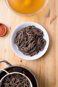 Nouilles soba dans une assiette blanche, avec un bol de bouillon et une passoire sur une table en bois.
