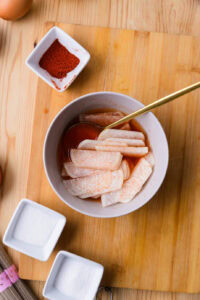 Tranches de radis marinées dans une sauce rouge, avec du sel et du piment en poudre sur une planche en bois.