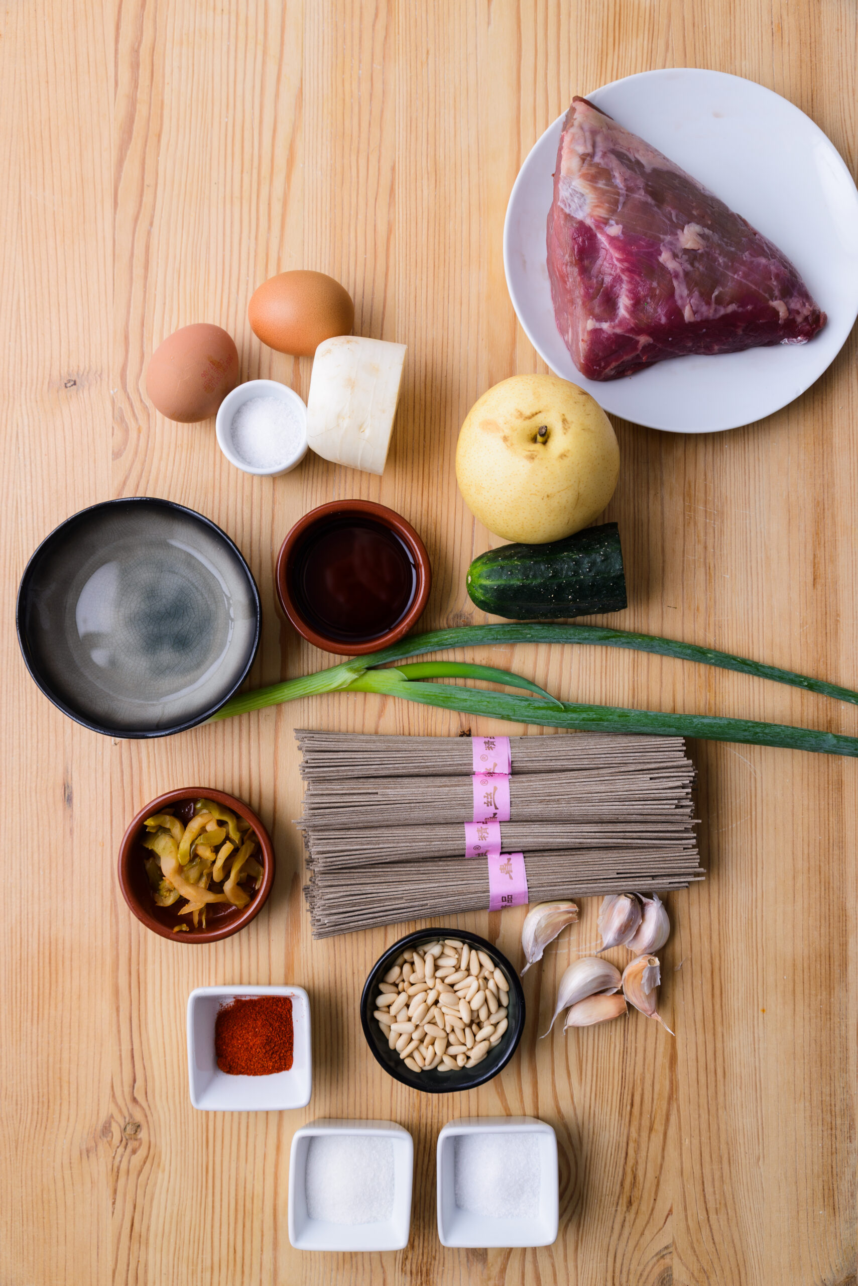 Ingrédients variés sur une table en bois : viande, nouilles, œufs, légumes, condiments et épices.