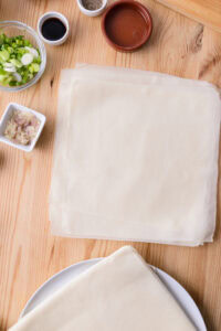 Feuilles de pâte à spring rolls sur une table en bois, entourées d’ingrédients hachés et de sauces en petits bols.