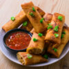 Assiette de lumpias dorés garnis d’oignons verts, accompagnés d’une sauce rouge pimentée.