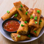 Assiette de lumpias dorés garnis d’oignons verts, accompagnés d’une sauce rouge pimentée.