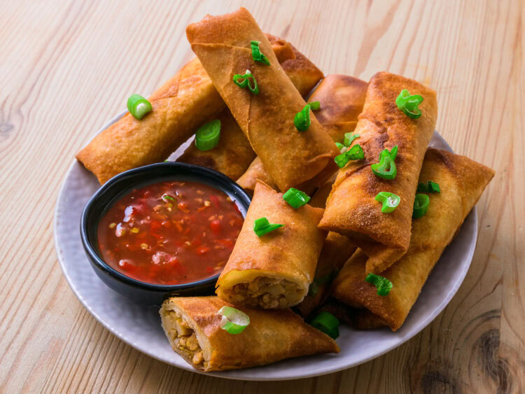 Assiette de lumpias dorés garnis d’oignons verts, accompagnés d’une sauce rouge pimentée.