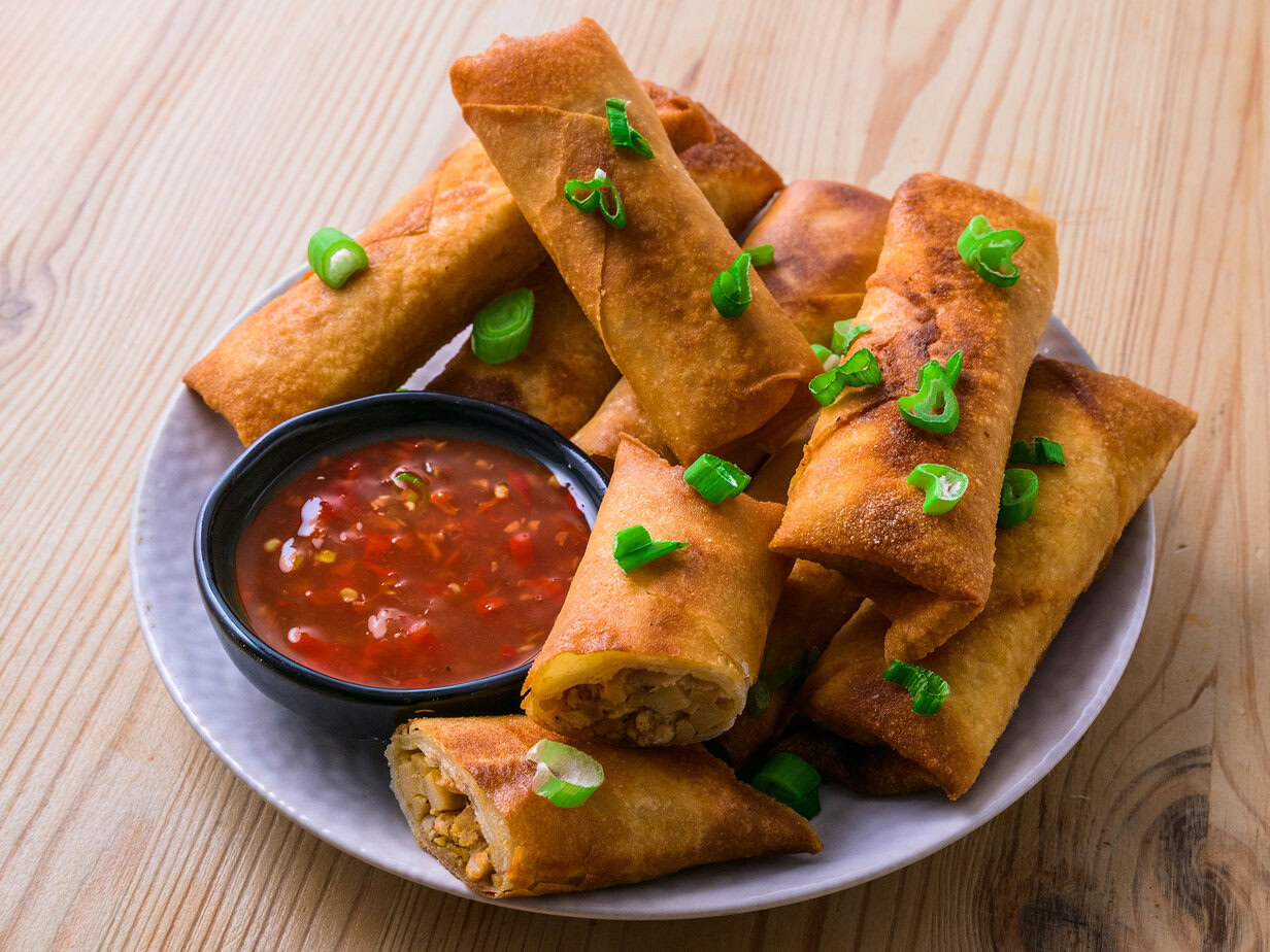Assiette de lumpias dorés garnis d’oignons verts, accompagnés d’une sauce rouge pimentée.