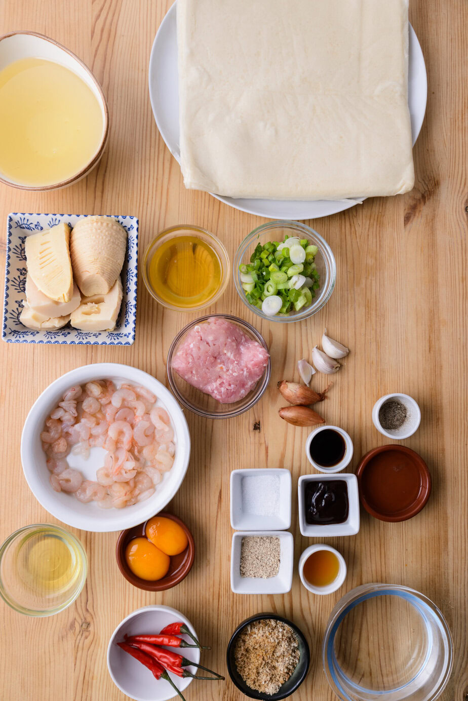 Ingrédients variés sur une table en bois : pâte, crevettes, viande, œufs, légumes, sauces et épices.