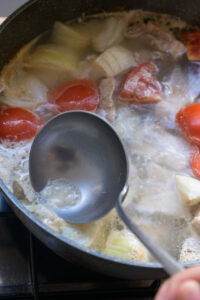 Loupe en métal écumant une soupe avec tomates, oignons et viande dans une casserole sur le feu.