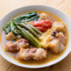 Bol de soupe avec morceaux de porc, légumes verts, gombo, tomate et bouillon sur une table en bois.