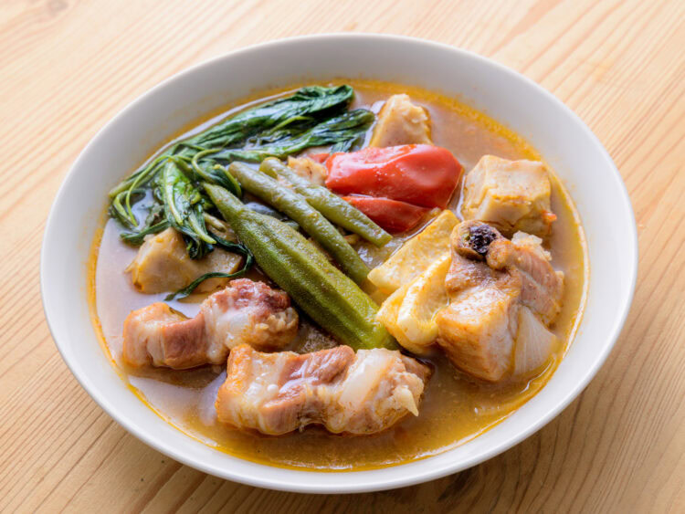 Bol de soupe avec morceaux de porc, légumes verts, gombo, tomate et bouillon sur une table en bois.
