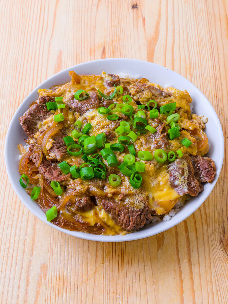Bol de gyudon japonais avec bœuf, œuf, oignons et ciboules sur du riz, servi sur une table en bois.