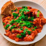 Poulet tikka masala garni de coriandre fraîche, servi avec des morceaux de pain naan dans une assiette blanche.