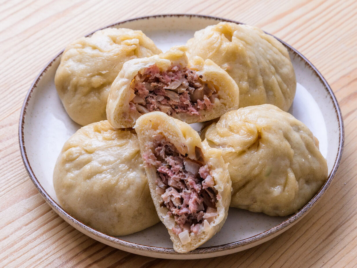 Cinq brioches vapeur farcies à la viande, dont une coupée en deux, servies sur une assiette.