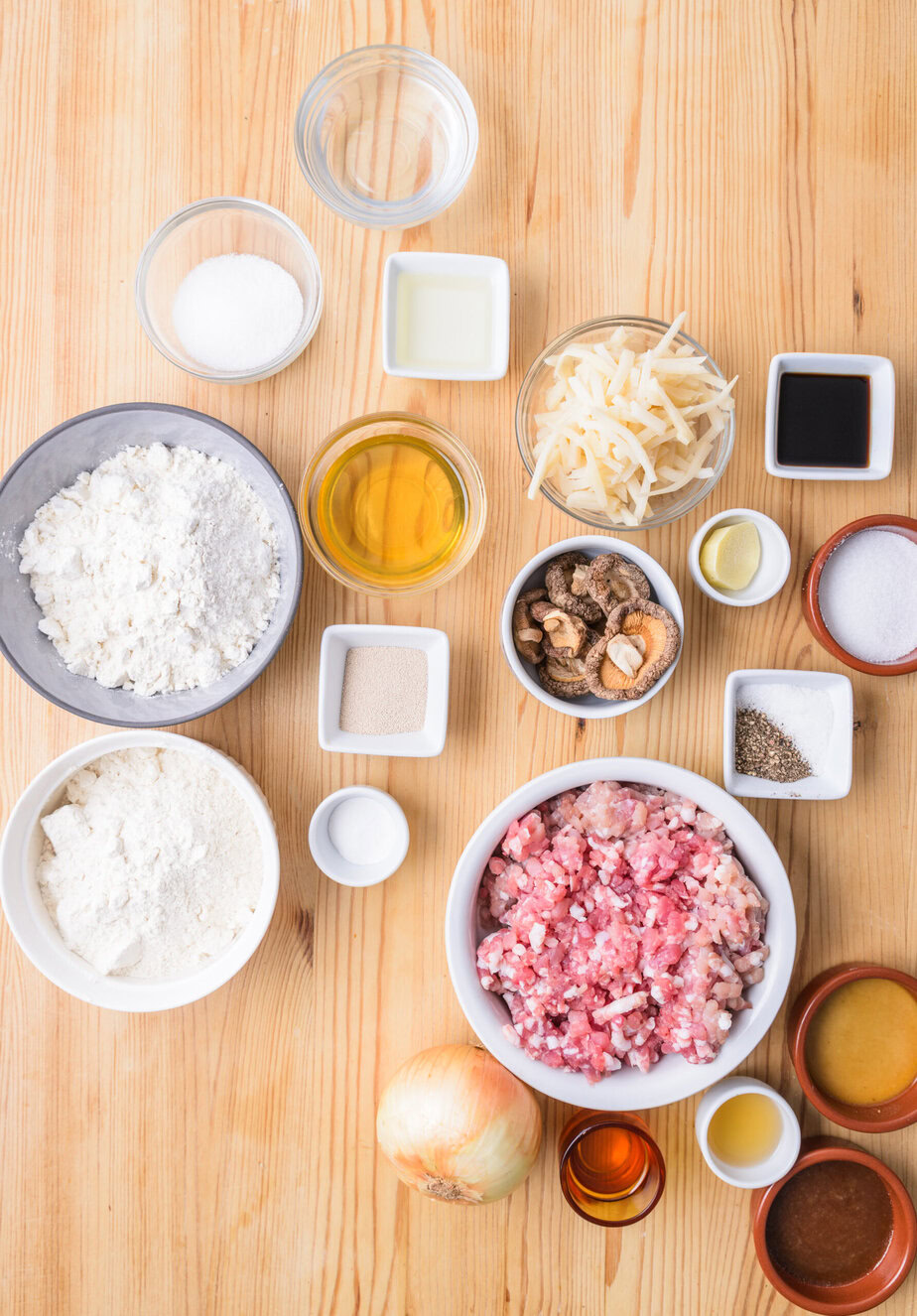 Ingrédients variés sur une table en bois : viande hachée, farine, oignon, épices, liquides et légumes râpés.