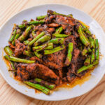 Porc sauté aux haricots verts et sauce épicée, servi dans une assiette blanche sur une table en bois.