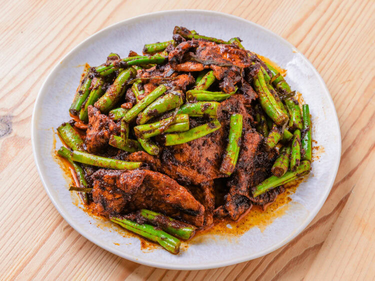 Porc sauté aux haricots verts et sauce épicée, servi dans une assiette blanche sur une table en bois.
