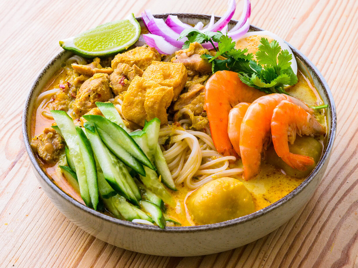 Bol de nouilles au curry avec crevettes, poulet, concombre, citron vert, coriandre et œuf sur une table en bois.
