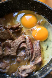 Bò Né - Pousser le bœuf d'un côté de la poêle. Casser les oeufs et le cuire au plat en gardant les jaunes coulants.