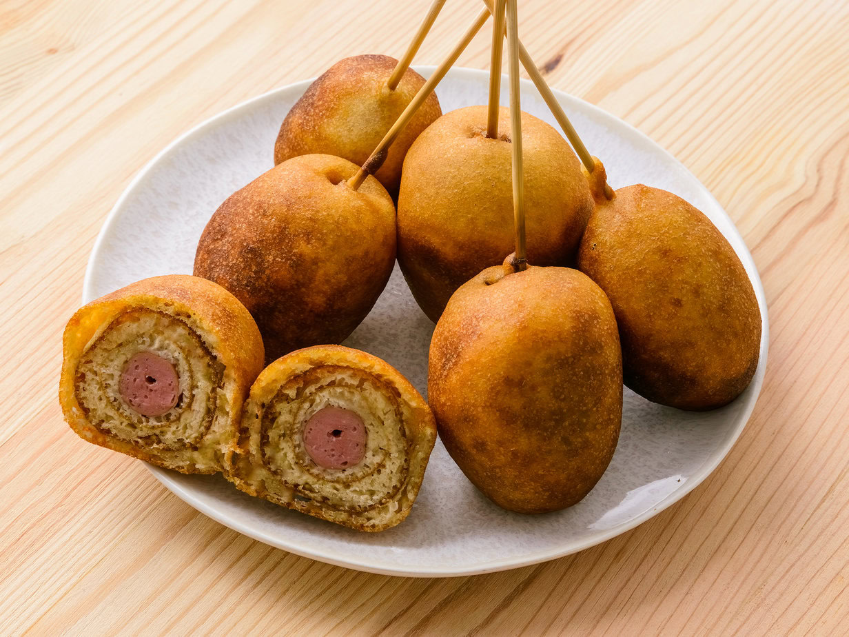 Corn dogs dorés sur une assiette blanche, dont un coupé en deux pour montrer la saucisse à l’intérieur.