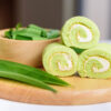 Roulés à la crème verts sur une planche en bois, avec des feuilles de pandan dans un bol.