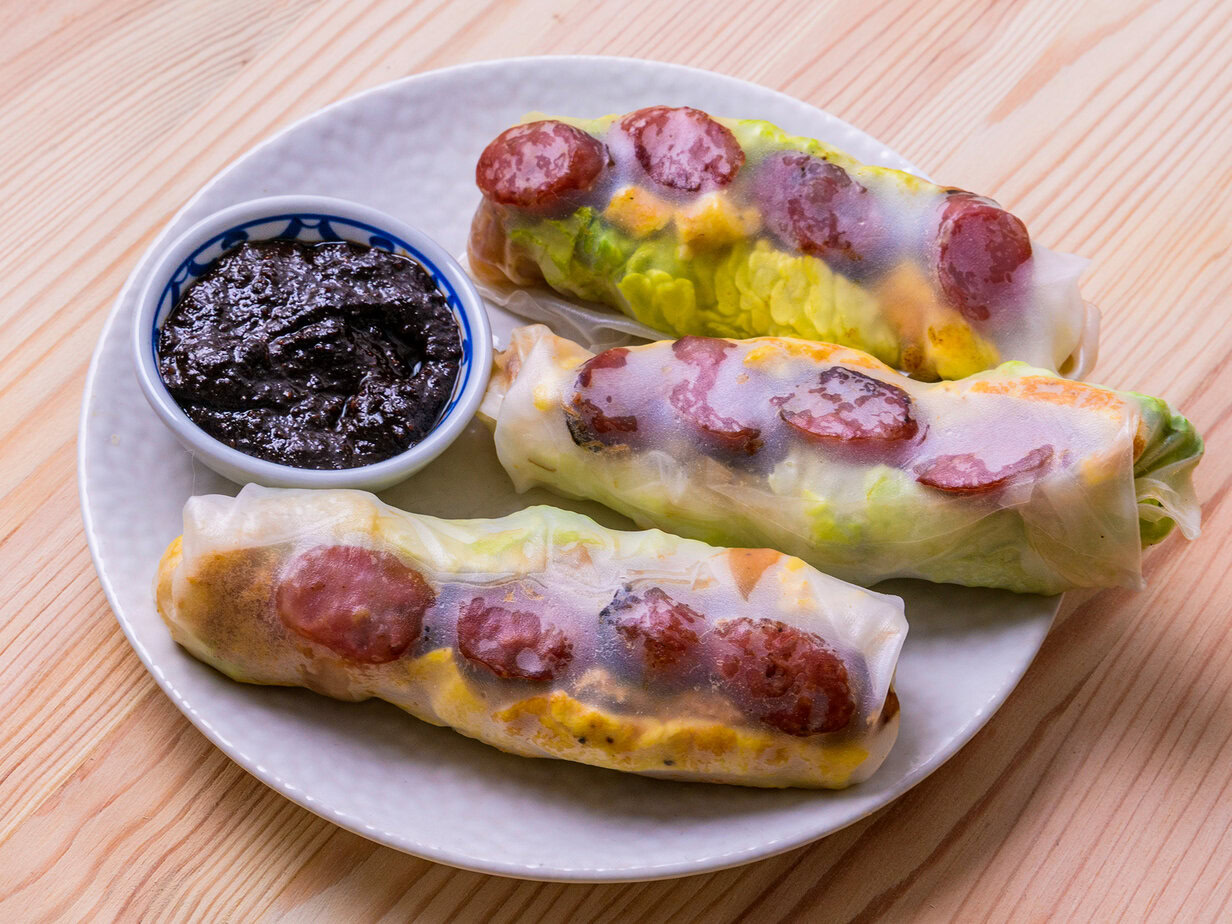 Trois rouleaux de printemps avec saucisse et légumes, accompagnés d'une sauce noire dans une petite coupelle.
