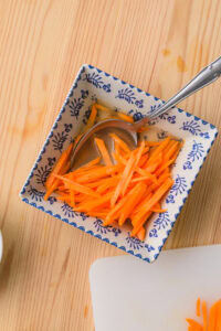Carottes râpées dans un bol carré décoré, avec une cuillère en métal.