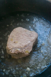 Un morceau de viande mijote dans une casserole avec des oignons émincés.