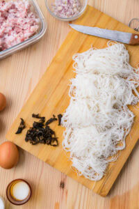 Nouilles de riz, champignons noirs, œufs, viande hachée et échalotes sur une planche à découper.