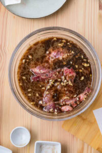Viande crue marinant dans une sauce brune avec des morceaux d’ail dans un bol en verre sur une table en bois.