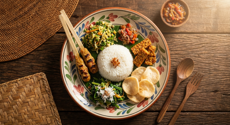 Assiette de riz blanc avec légumes, brochettes, tempeh, chips et sauce, sur une table en bois.