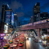 Rue animée de nuit à Bangkok, avec circulation dense, gratte-ciel et métro aérien.