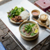 Repas vietnamien avec pho, boulettes, vermicelles, herbes fraîches, sauce, café, sur une table de rue.