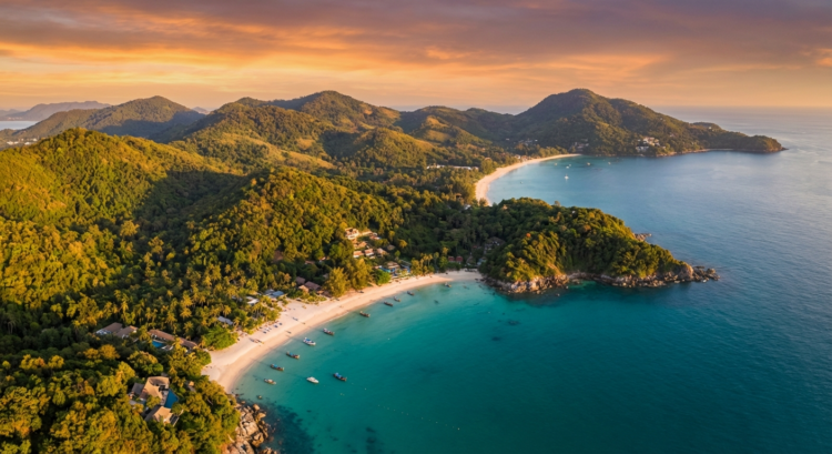 Plage de sable blanc bordée de forêt tropicale et mer turquoise, vue aérienne au coucher du soleil.