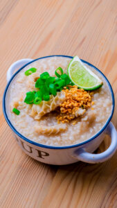 Bol de soupe de riz garni de tripes, ciboulette, ail frit et quartier de citron vert sur une table en bois.