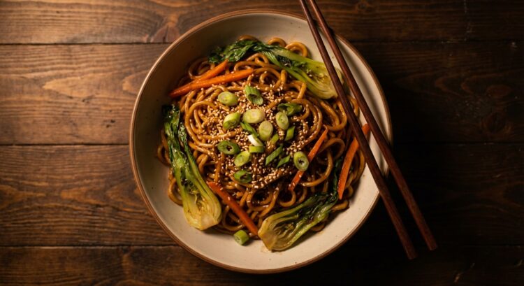 Bol de nouilles sautées aux légumes, garni de graines de sésame et oignons verts, avec baguettes.
