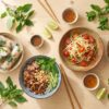Assortiment de plats vietnamiens : rouleaux de printemps, salade, nouilles, herbes fraîches et sauces sur table en bois.