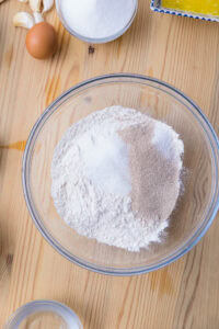 Bol de farine, sucre et levure sur une table en bois, avec œuf, ail et beurre fondu autour.