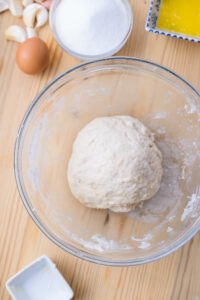 Pâte à pain en boule dans un saladier en verre, avec œuf, sucre et beurre autour.