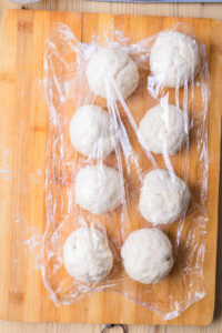 Boules de pâte levée couvertes de film plastique sur une planche en bois.