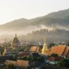 Ville asiatique avec temples dorés au pied de collines verdoyantes enveloppées de brume matinale.