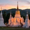 Temple bouddhiste blanc et doré au coucher du soleil, devant des collines boisées.