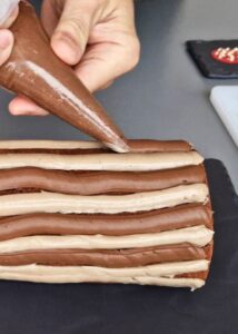 Bûche sans gluten chocolat et châtaigne - Retaille les deux extrémités de la bûche pour révéler une garniture uniforme. Déplace sur un plateau à gâteau ou une assiette. Poche des lignes alternées de crèmes pâtissières à la châtaigne et au chocolat jusqu’à couvrir toute la surface de la bûche.