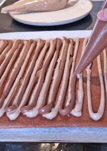 Bûche sans gluten chocolat et châtaigne - Coupe les bords secs de la génoise. La génoise doit mesurer 28 cm de large et 38 cm de long. Pose la génoise sur du papier cuisson, puis poche les crèmes pâtissières à la châtaigne et au chocolat en alternance pour couvrir la surface, en laissant 1 cm de marge par rapport aux bords.